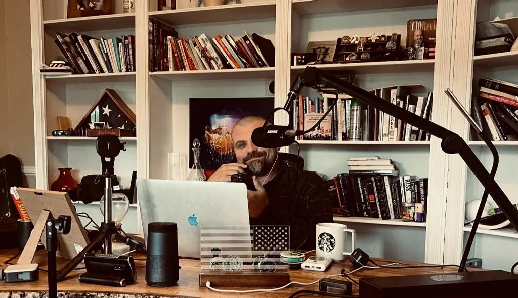 Daniel recording a podcast at a desk with a microphone, bookshelves, and American flags.